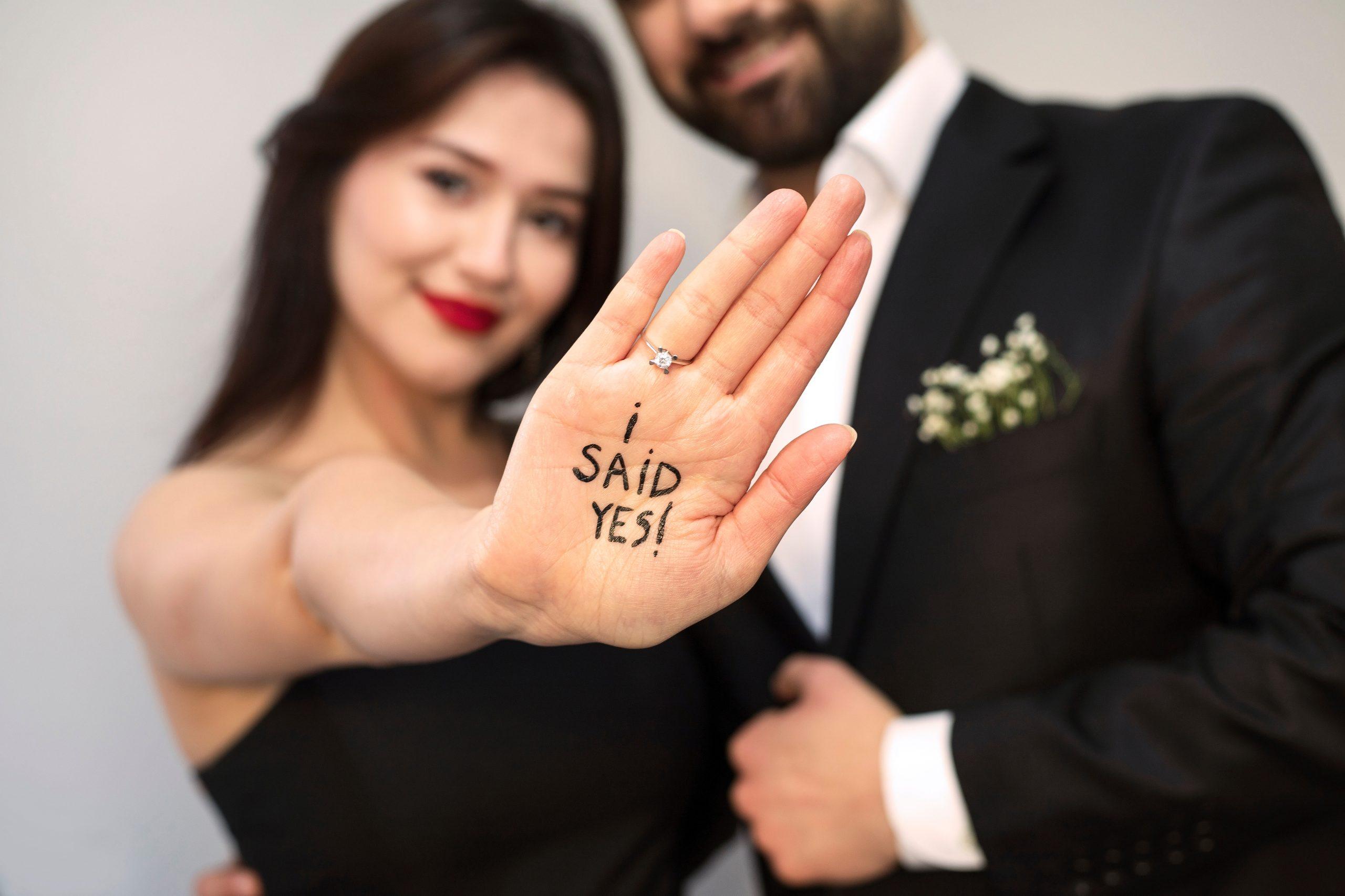 A young romantic couple standing together in black tie clothing. The woman holds our her hand with the palm facing the camera which displays a diamond engagement ring and writing on her palm in black that says "i SAiD YES!"
