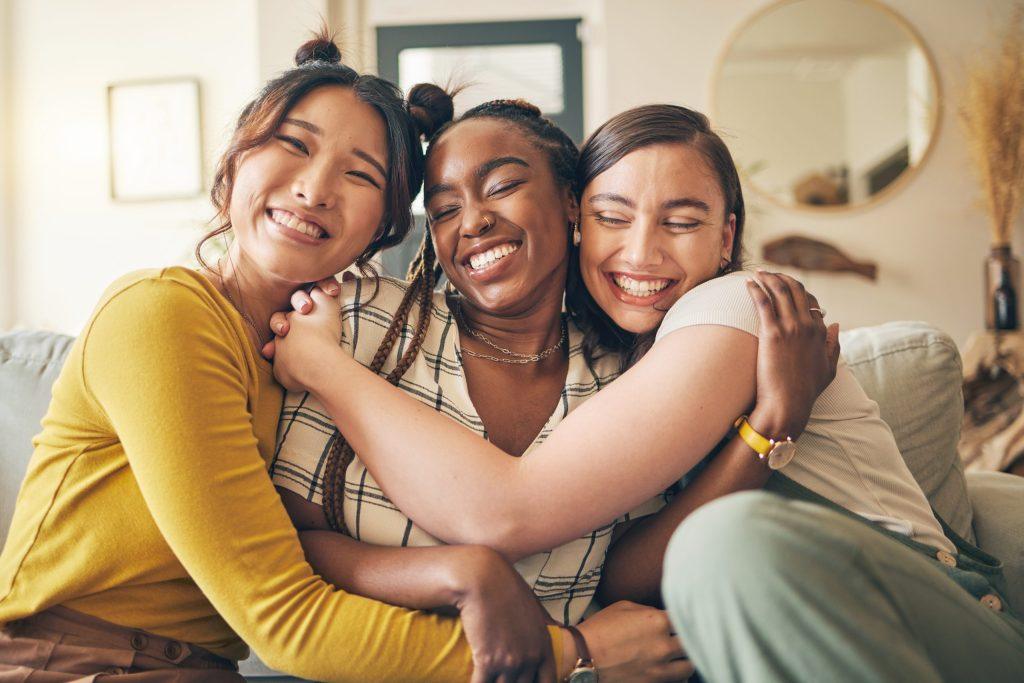 Portrait of a group of women, friends hug on sofa with smile and bonding in living room together in embrace. Hug, love and friendship, girls on couch with diversity, pride and people in home with fun