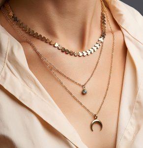 Cropped close-up portrait of a woman, demonstrating gold necklaces with pendants in a beige shirt. Set of three different chains. One necklace has a crescent moon pendant.
