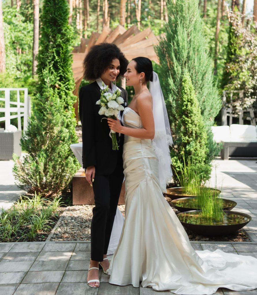 Cheerful interracial lesbian couple with bouquet looking at each other during wedding outdoors.